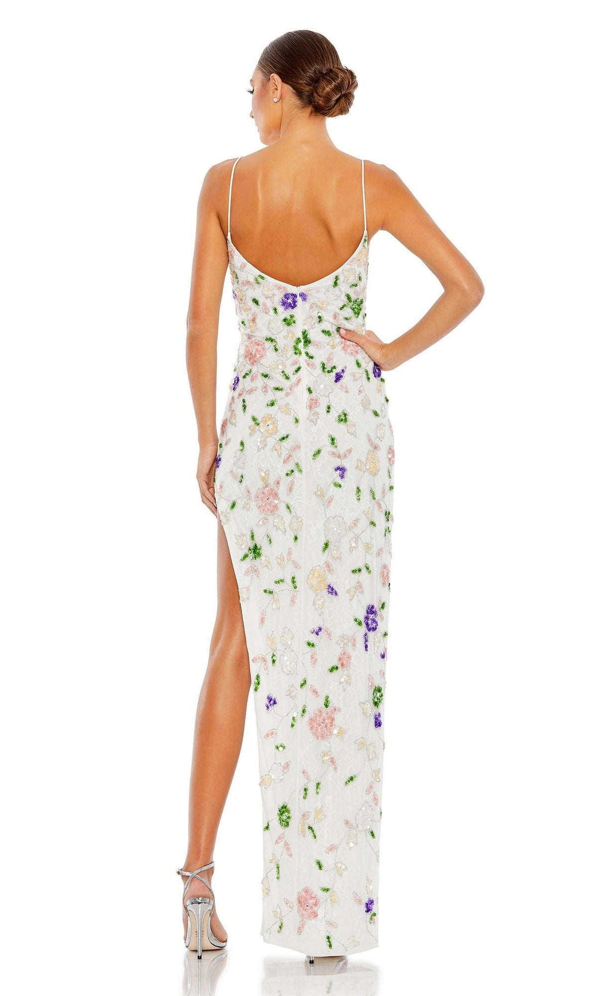 A woman faces away, wearing the Long Formal Dress 10893 by Mac Duggal—a white, floor-length fitted gown with floral patterns, spaghetti straps, a high slit—and silver high-heeled sandals. Her hair is styled in a neat bun.